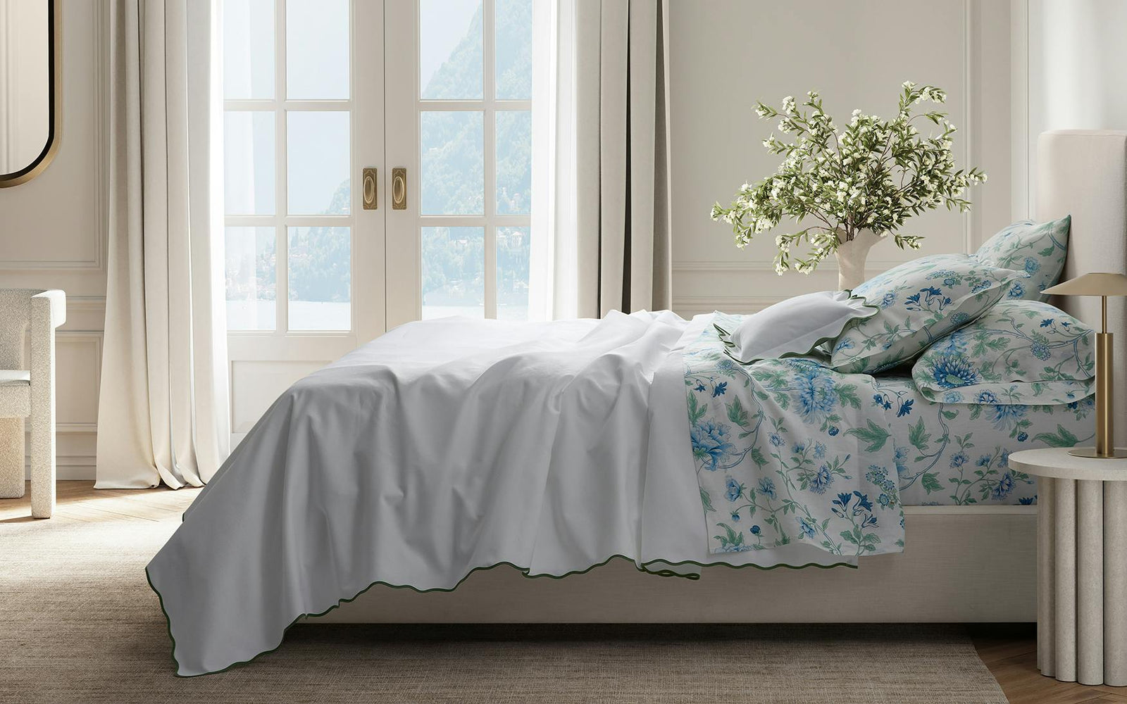 A neatly made bed with white and floral bedding in a bright room with large windows, curtains, and a plant.