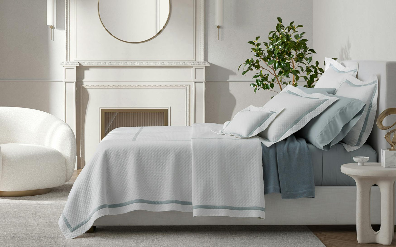 A neatly made bed with white bedding, a round mirror above a fireplace, a plant, and a white chair in a bright bedroom.