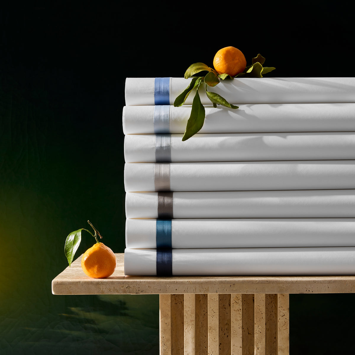 A stack of folded Matouk Lowell Egyptian cotton percale sheets, topped with tangerines and leaves on a stone table.