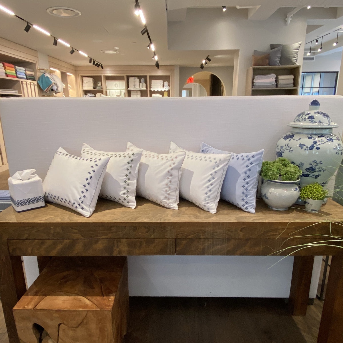 Five white decorative pillows, inspired by Matouk Casablanca Bedding, sit on a table with jars and green plants.