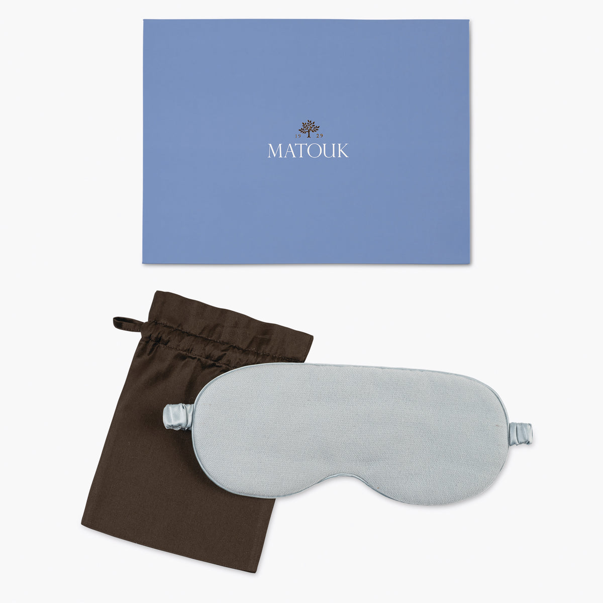 A light blue silk sleep mask, a brown drawstring pouch, and a blue Matouk box are arranged on a white background.
