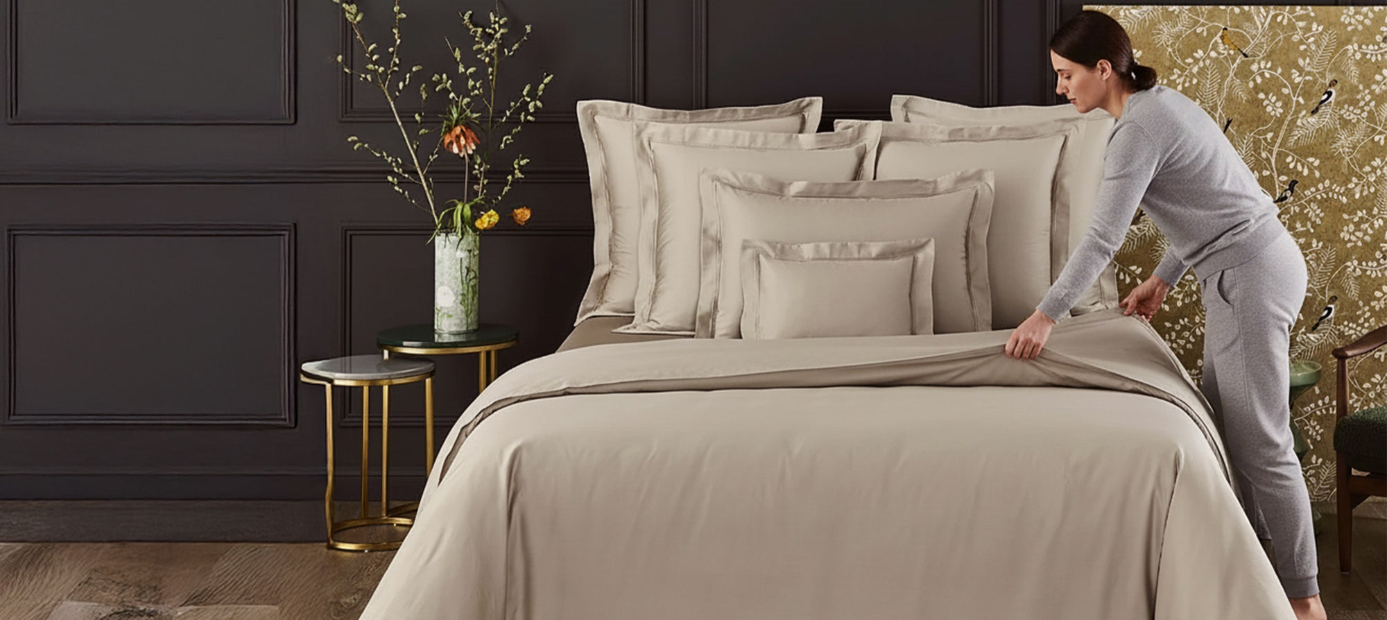 Woman arranging bedding in a bedroom with dark paneled walls and decorative elements.