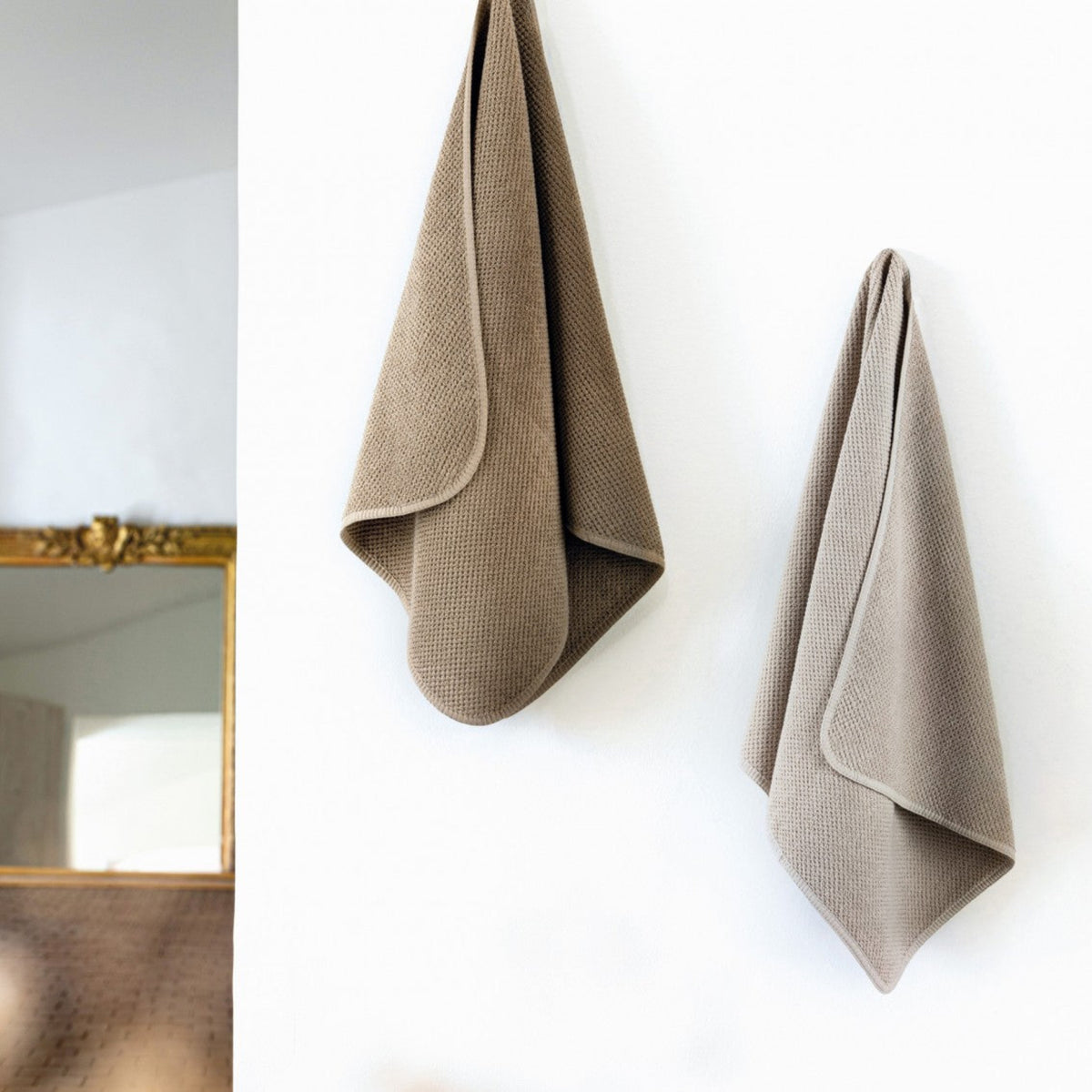 Two beige towels hanging on a white wall with a gold-framed mirror.