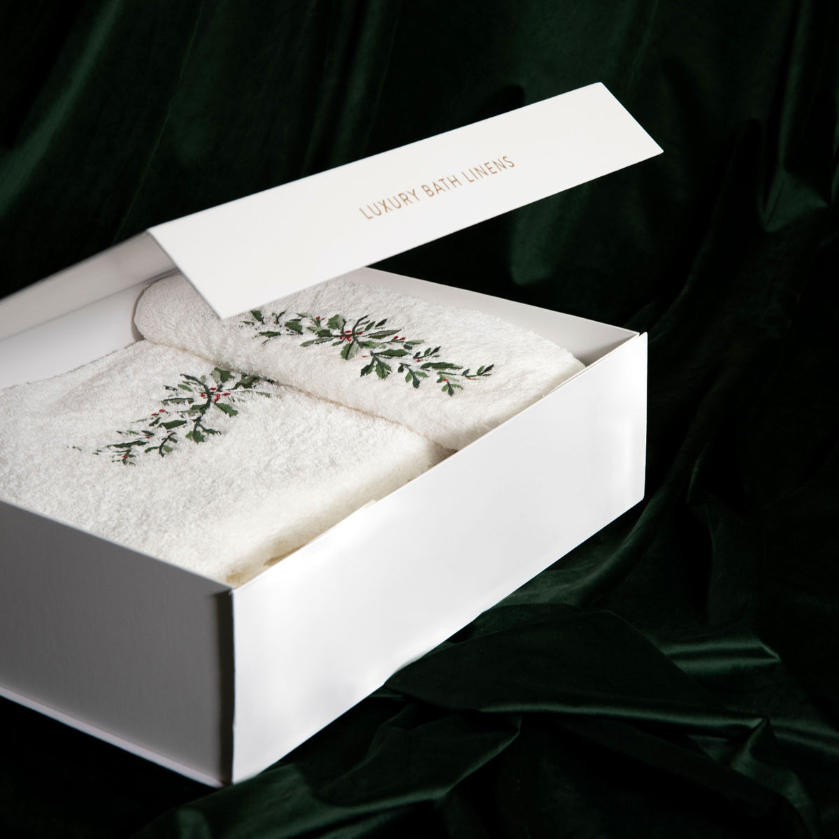 White gift box labeled Luxury Bath Linens containing two Graccioza Festive Towels with embroidered holly sprigs.