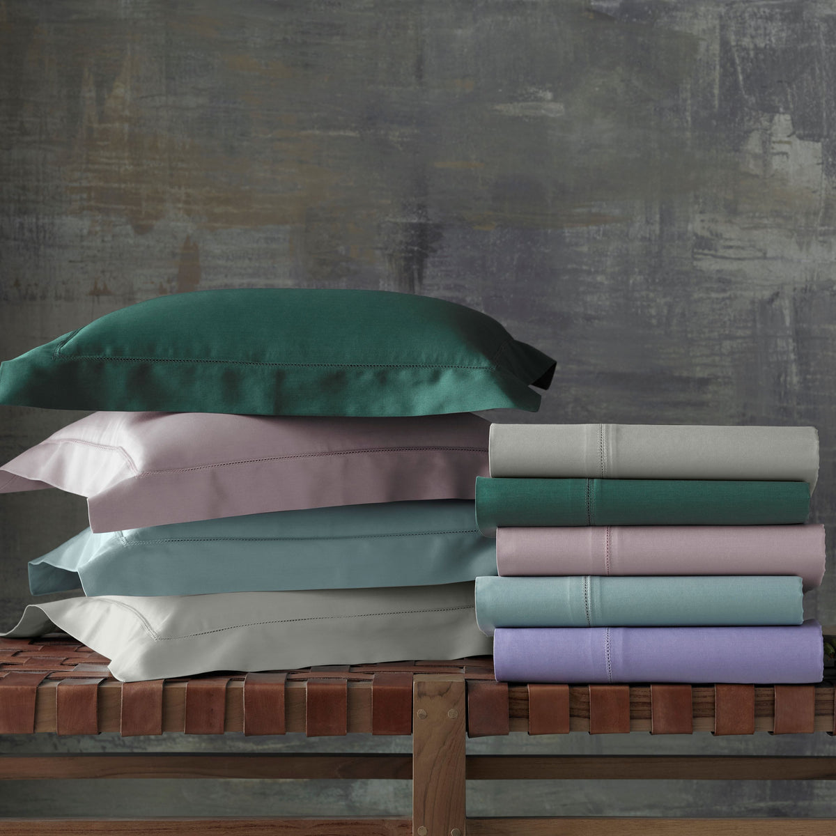 Stacked folded Egyptian cotton bedding and pillowcases in pastel colors on a wooden bench against a textured wall. Folded stack colors are Beach, Forest, Dusty Rose, Irish Winter Green and Violet (discontinued).