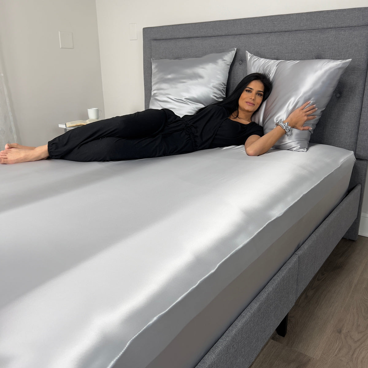 Woman lying on silk fitted sheet in Silver and matching pillows on her back, wearing black outfit