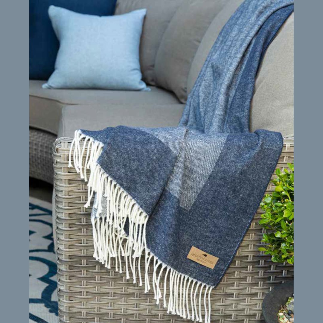 Lands Downunder Border Herringbone throw blanket with white fringe draped over a wicker chair next to a gray couch with blue and gray cushions.