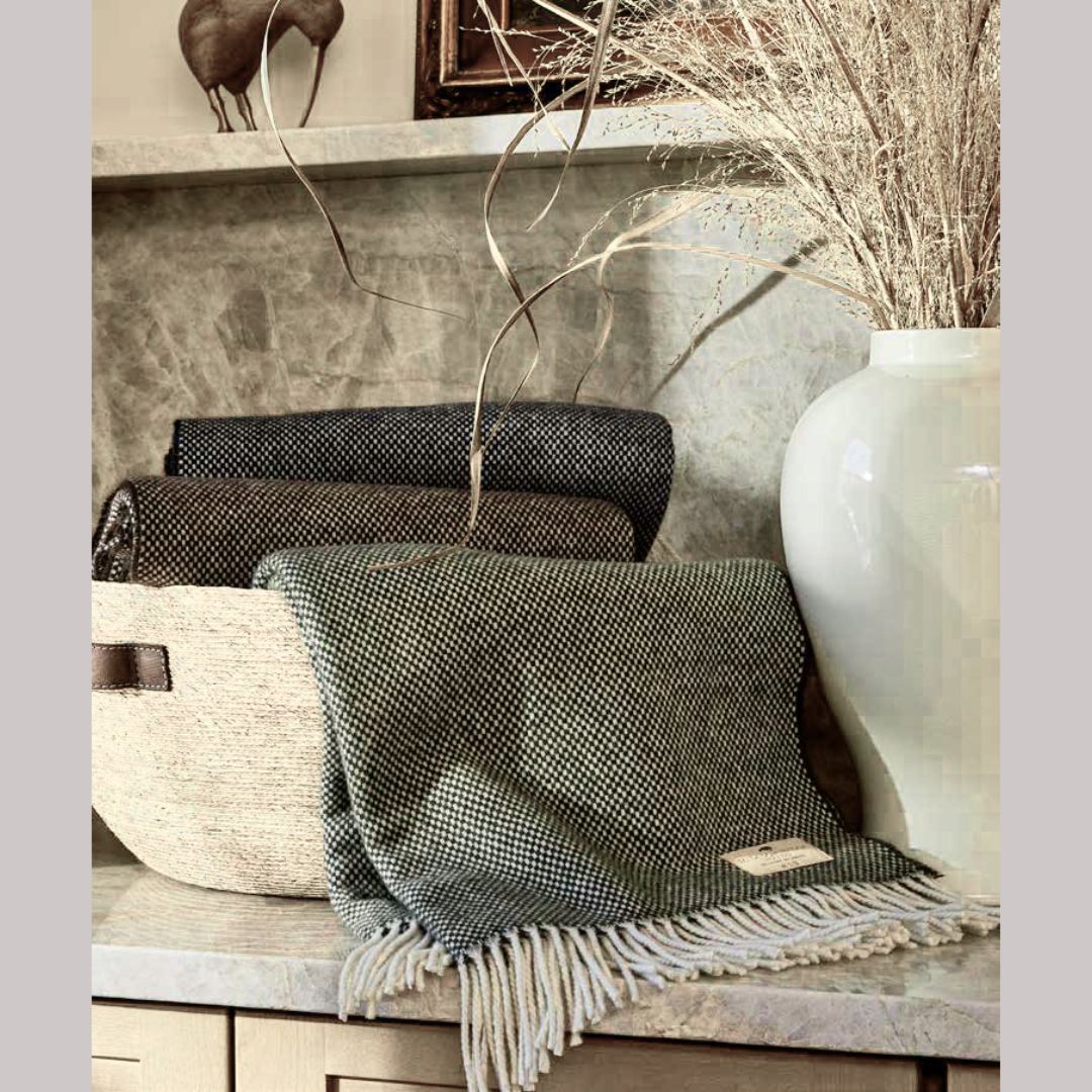 A woven basket holds a folded cashmere throw beside a white vase with dried grass on a marble countertop.