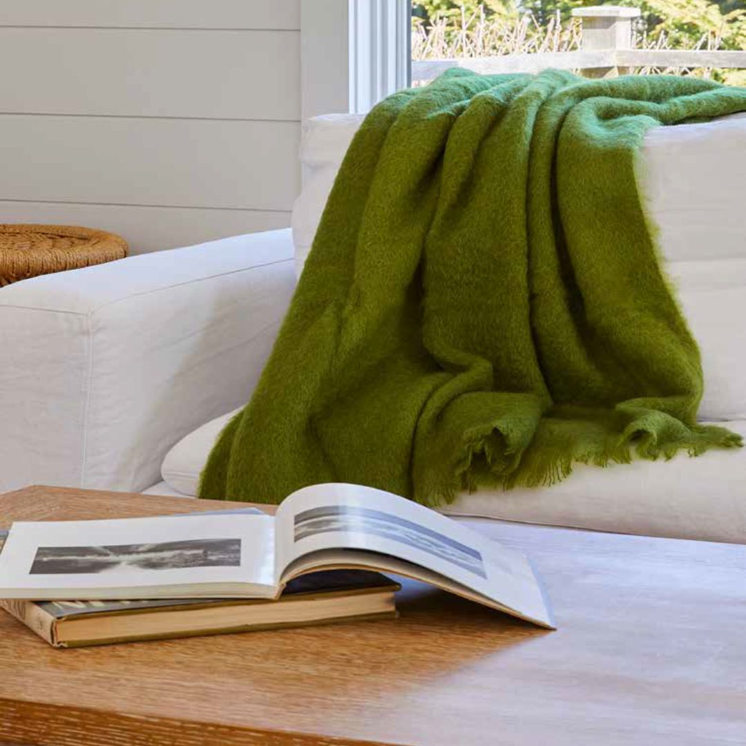 Lands Downunder New Zealand Mohair throw in Fern draped over a white sofa with an open book on a wooden table in the foreground.