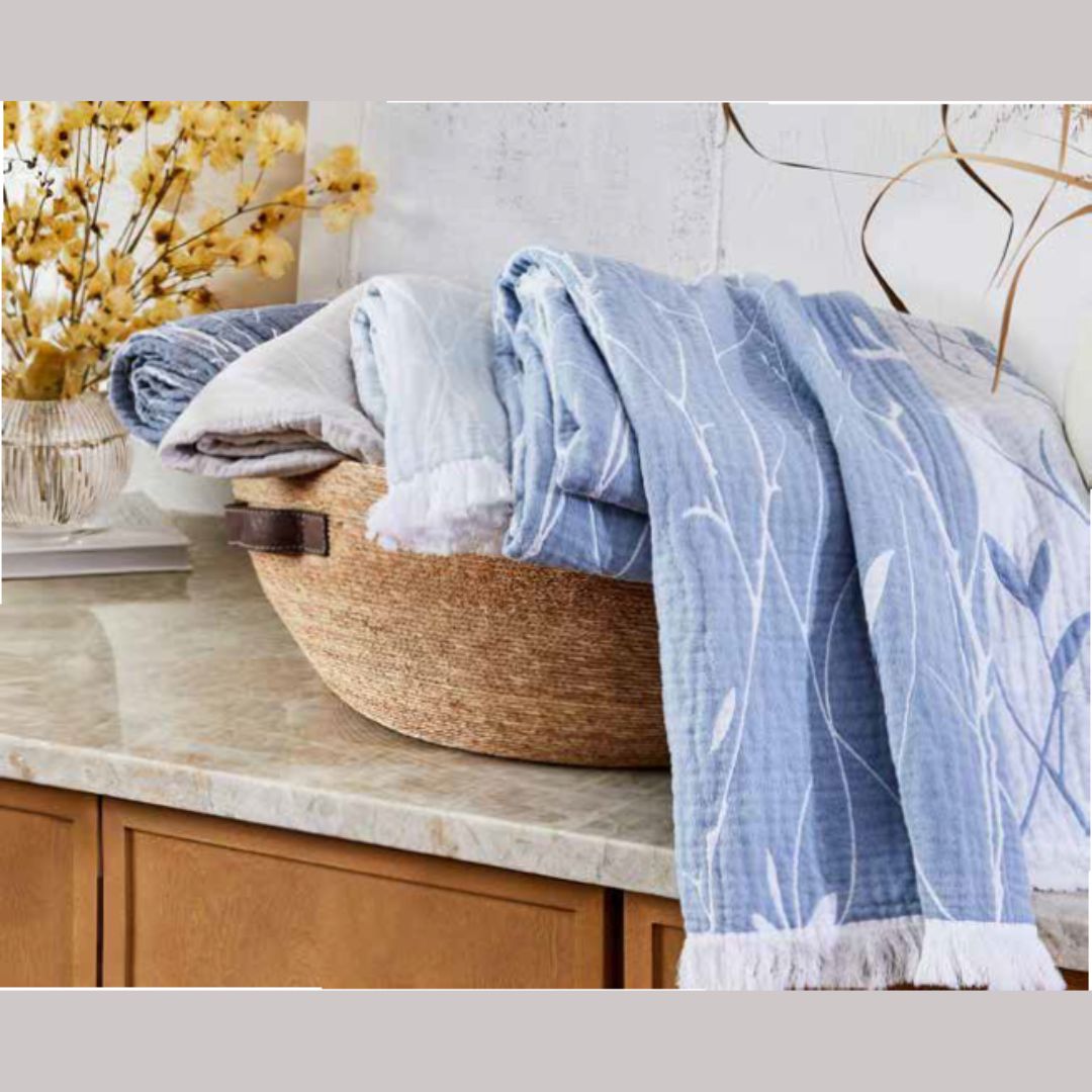 A woven basket with a Lands Downunder Willow throw blanket sits on a marble countertop next to dried yellow flowers.