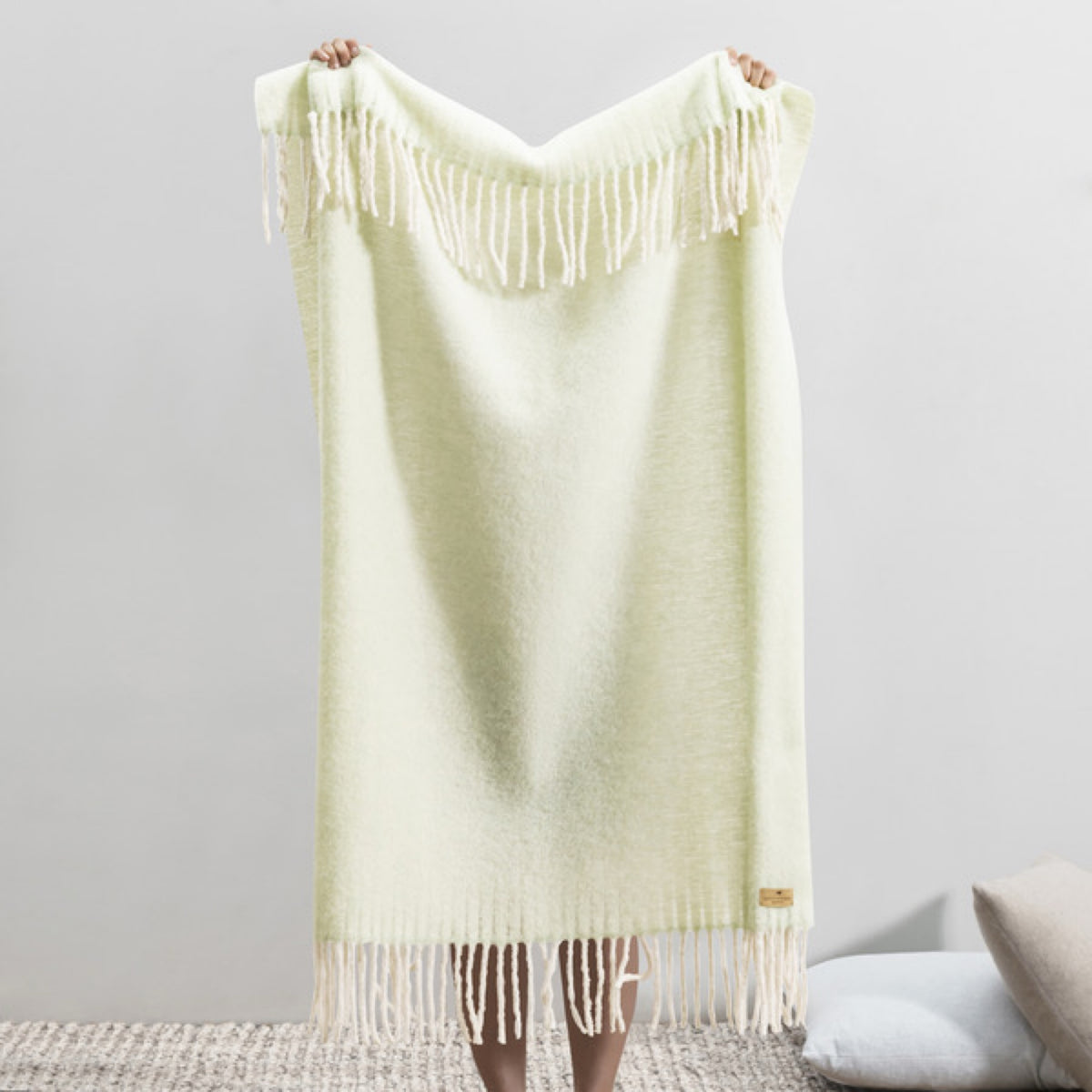 Person holding up a Lands Downunder Andes Throw in Celadon color with white fringe, standing indoors on a textured rug
