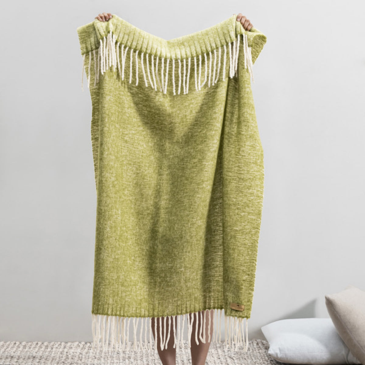 Person holding up a Lands Downunder Andes Throw in Fern color with white fringe, standing indoors on a textured rug