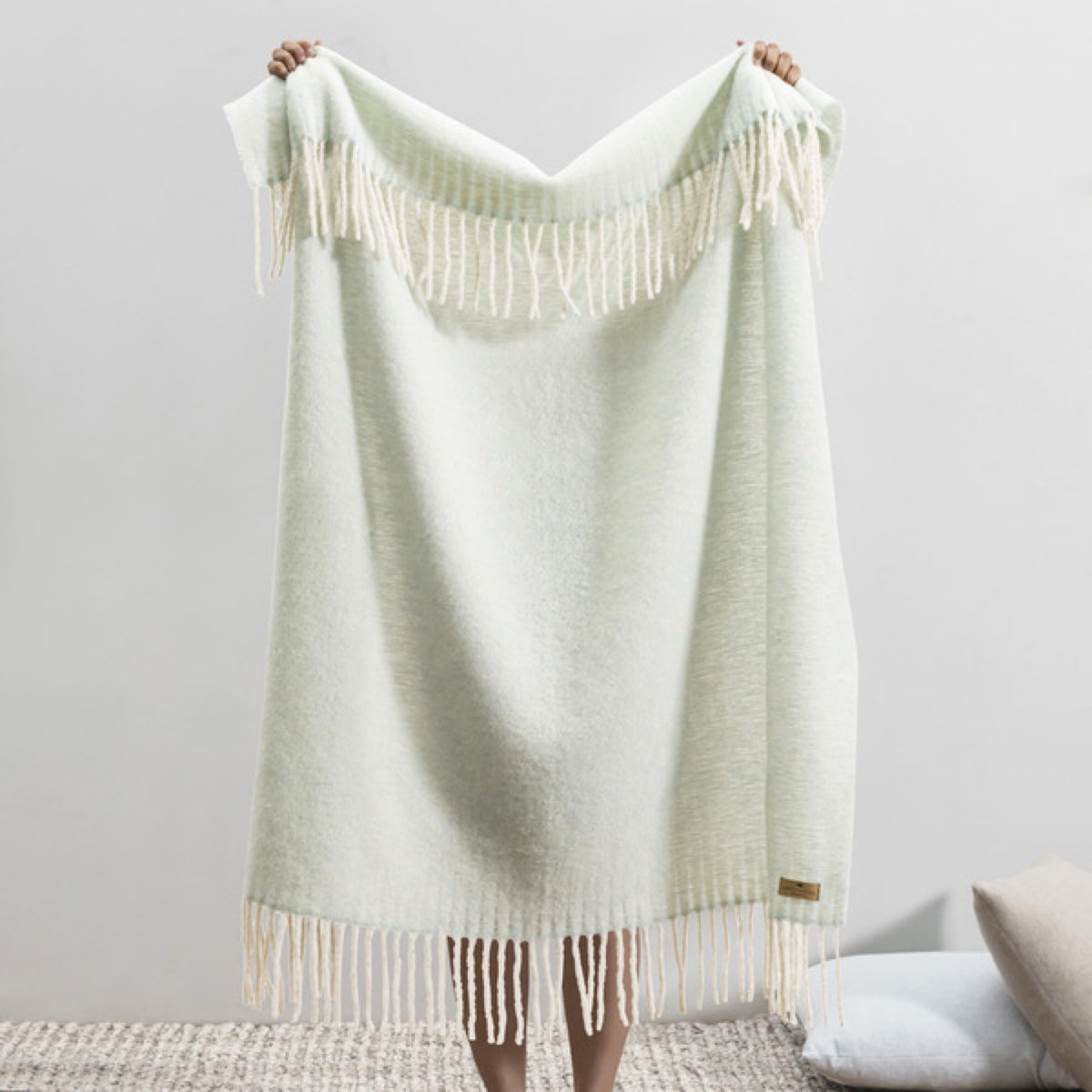 Person holding up a Lands Downunder Andes Throw in Glacier color with white fringe, standing indoors on a textured rug