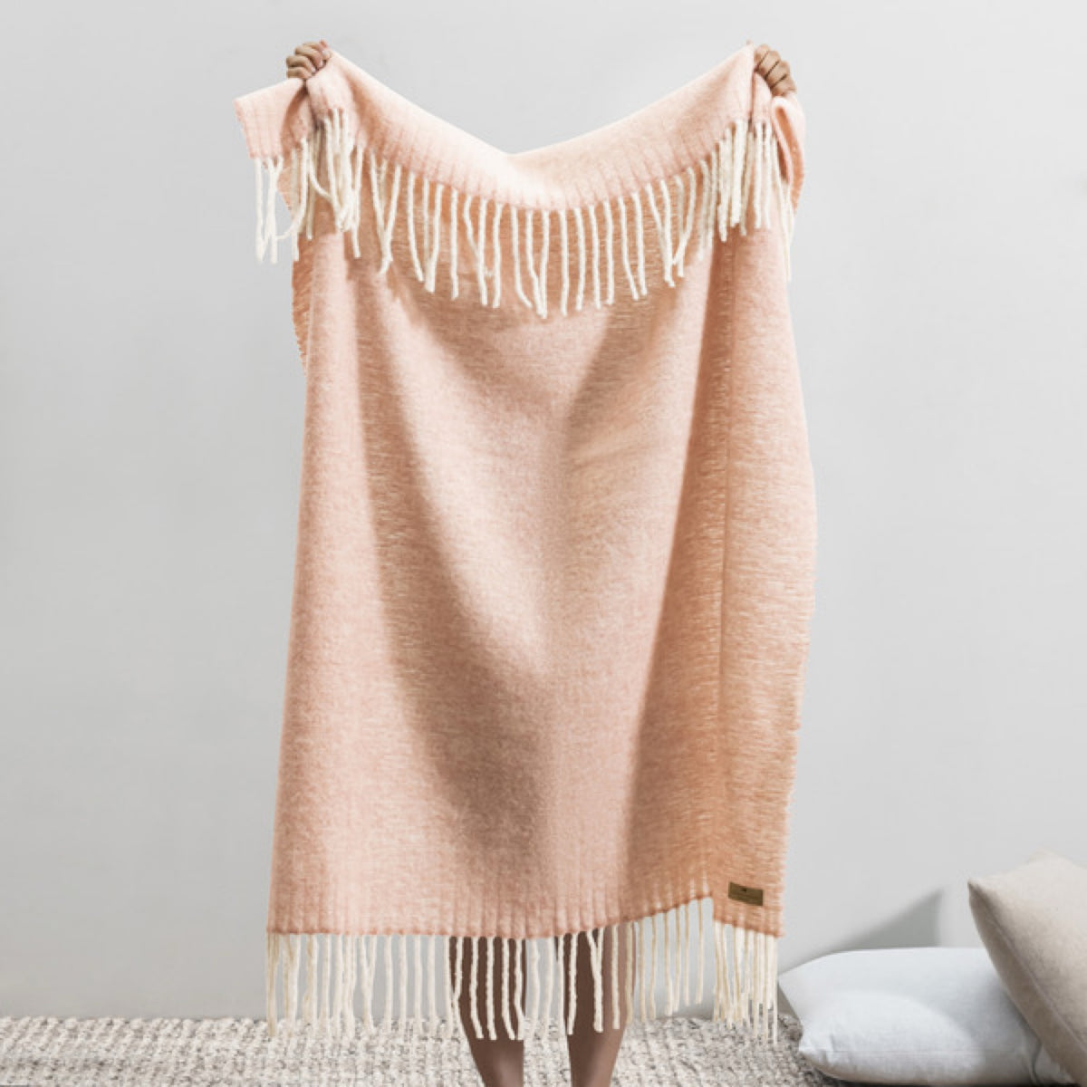 Person holding up a Lands Downunder Andes Throw in Rose Quartz color with white fringe, standing indoors on a textured rug