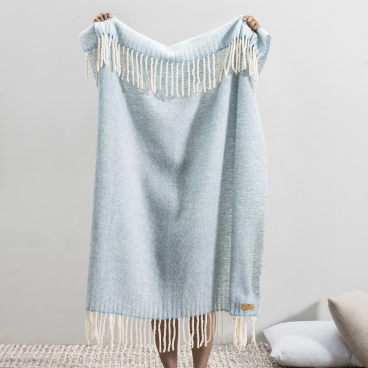 Person holding up a Lands Downunder Andes Throw in Sky Blue color with white fringe, standing indoors on a textured rug