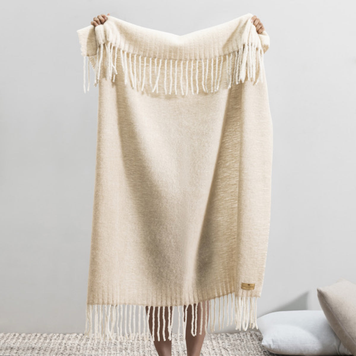 Person holding up a Lands Downunder Andes Throw in Truffle color with white fringe, standing indoors on a textured rug
