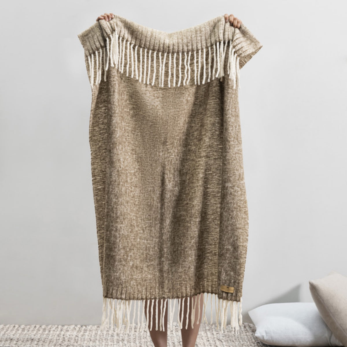 Person holding up a Lands Downunder Andes Throw in Walnut color with white fringe, standing indoors on a textured rug