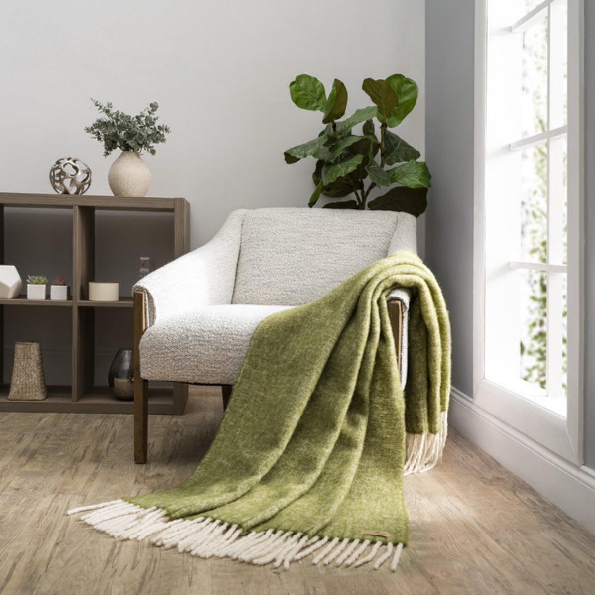 A Lands Downunder Andes Throw in Fern color draped over a light armchair by a window adds cozy home decor