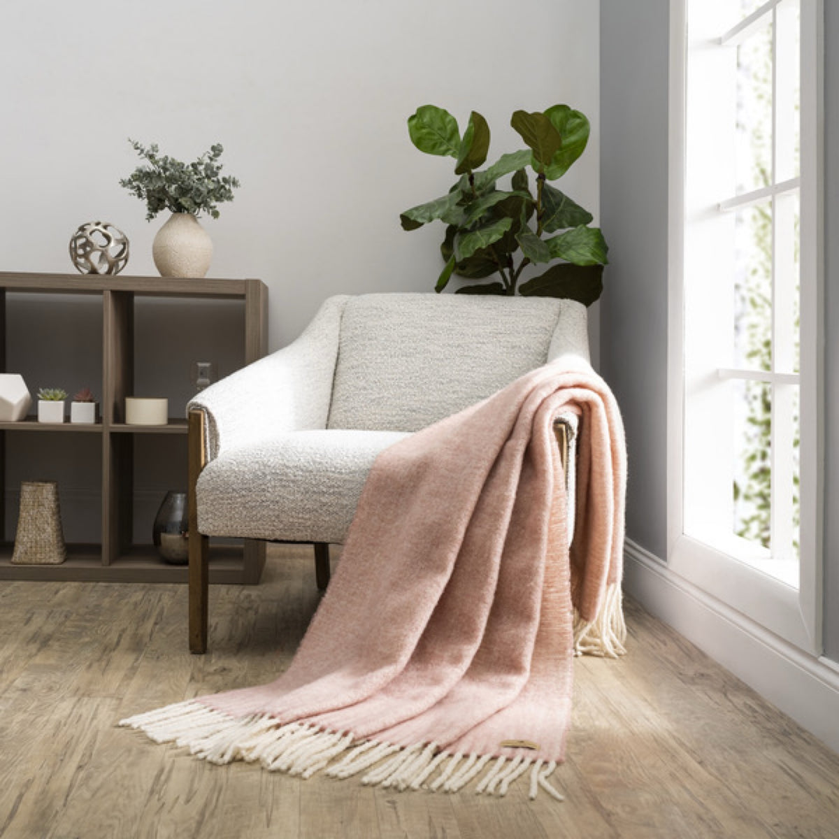 A Lands Downunder Andes Throw in Rose Quartz color draped over a light armchair by a window adds cozy home decor