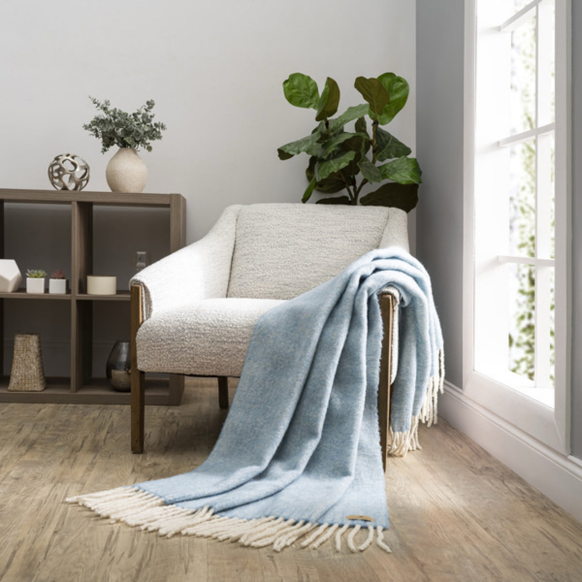 A Lands Downunder Andes Throw in Sky Blue color draped over a light armchair by a window adds cozy home decor