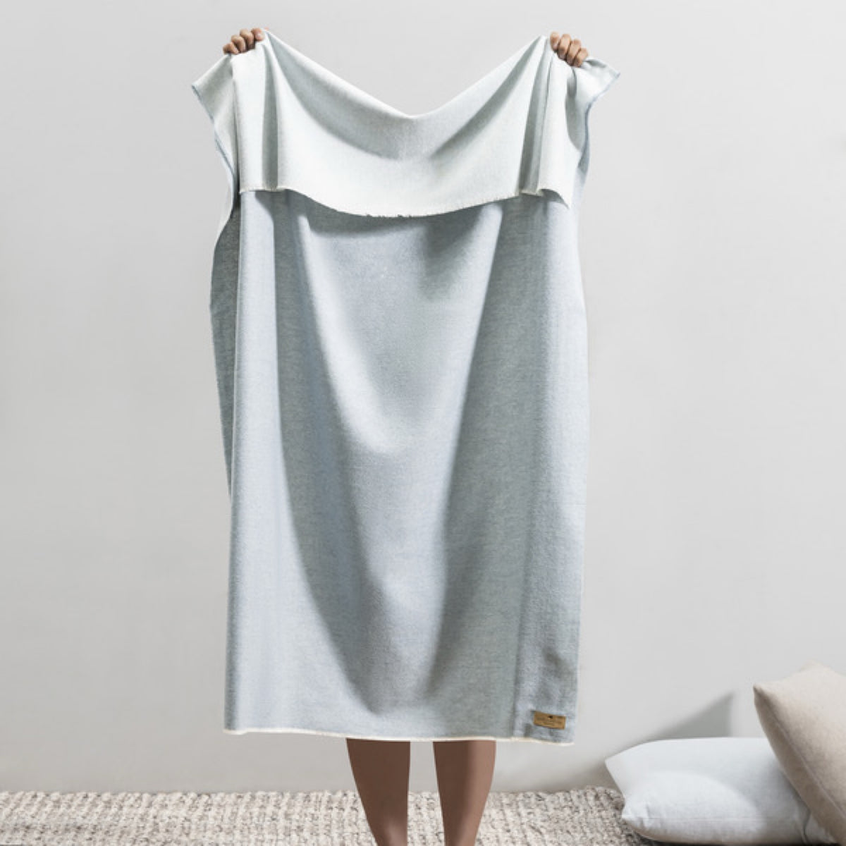 A person stands barefoot on a rug, holding up a Lands Downunder Aspen Peaks Throw in Crystal Blue color