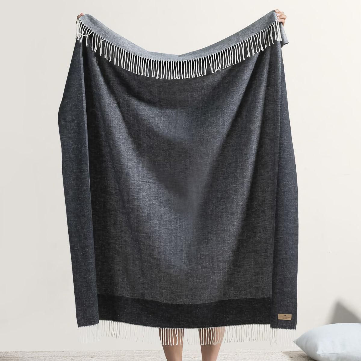 Person holding up a luxury throw blanket, the Border Herringbone Throw, against a plain light-colored wall and floor.