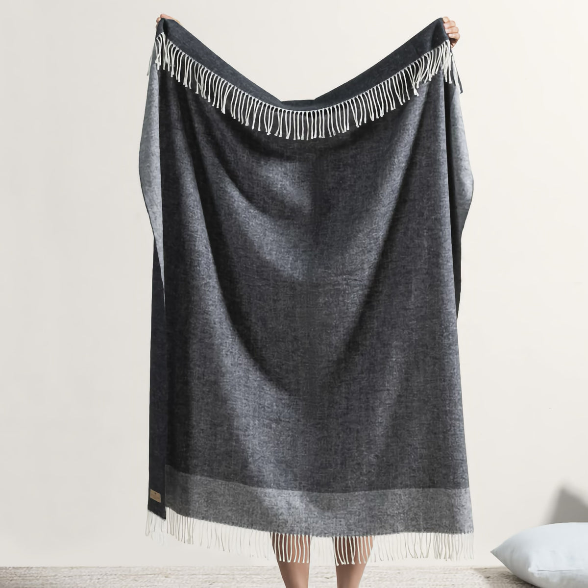A person holds up a luxury throw blanket, the Border Herringbone Throw, standing on a light floor with a pillow nearby.