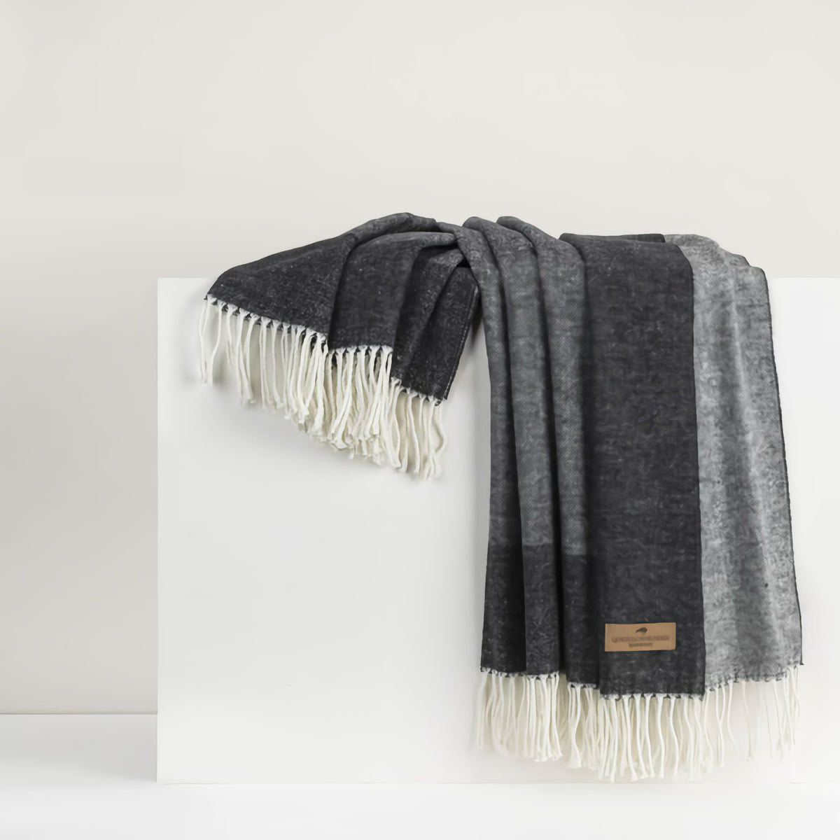 A dark gray luxury throw blanket with white fringe is draped over a white surface against a light background.
