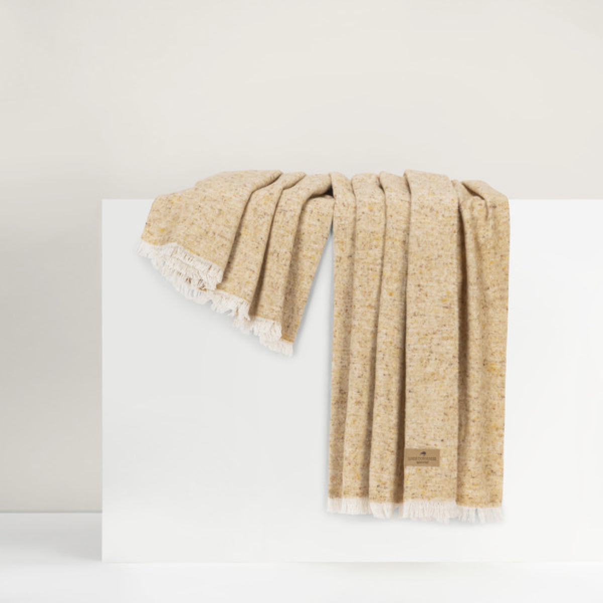 Lands Downunder Cinder Mist Throw in Gold color with fringed edges is draped over a white surface against a neutral background