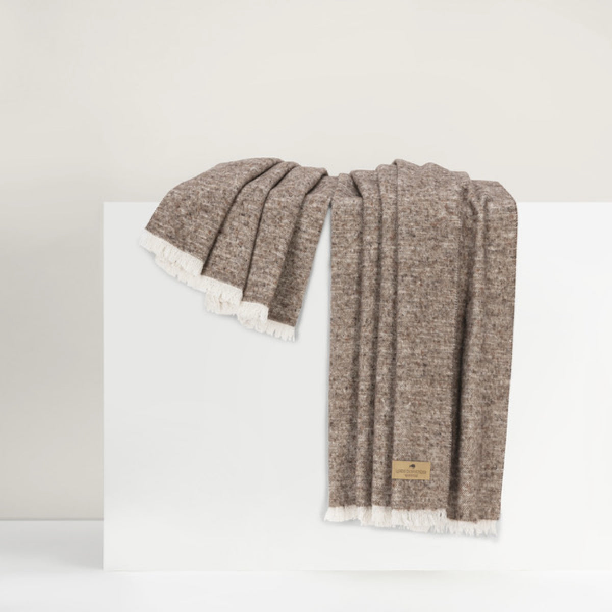 Lands Downunder Cinder Mist Throw in Mocha color with fringed edges is draped over a white surface against a neutral background