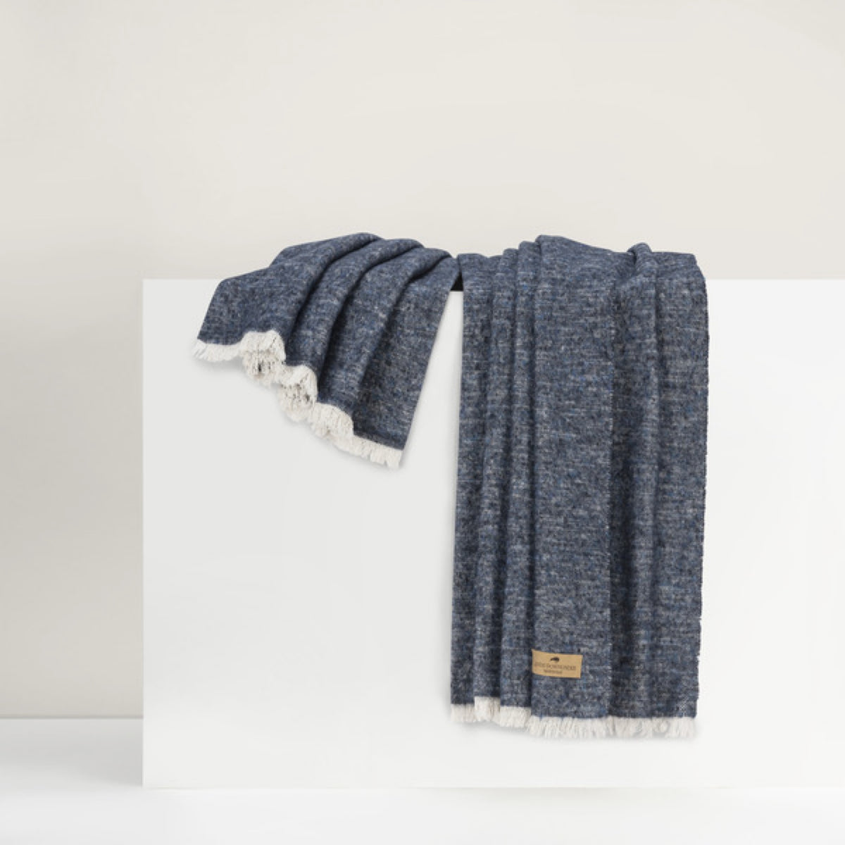 Lands Downunder Cinder Mist Throw in Navy color with fringed edges is draped over a white surface against a neutral background
