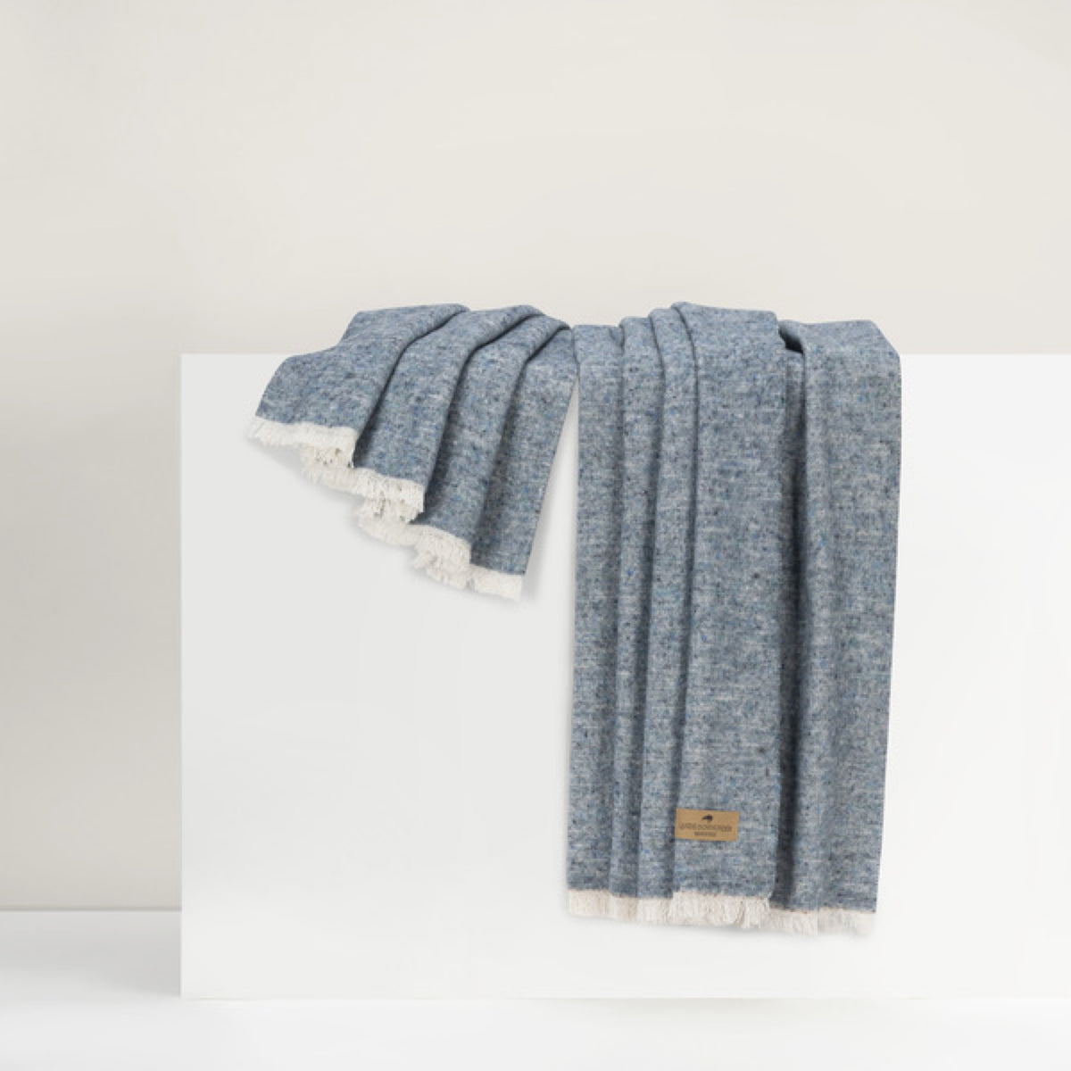 Lands Downunder Cinder Mist Throw in Ocean color with fringed edges is draped over a white surface against a neutral background
