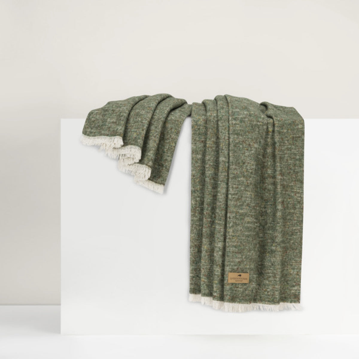 Lands Downunder Cinder Mist Throw in Olive color with fringed edges is draped over a white surface against a neutral background