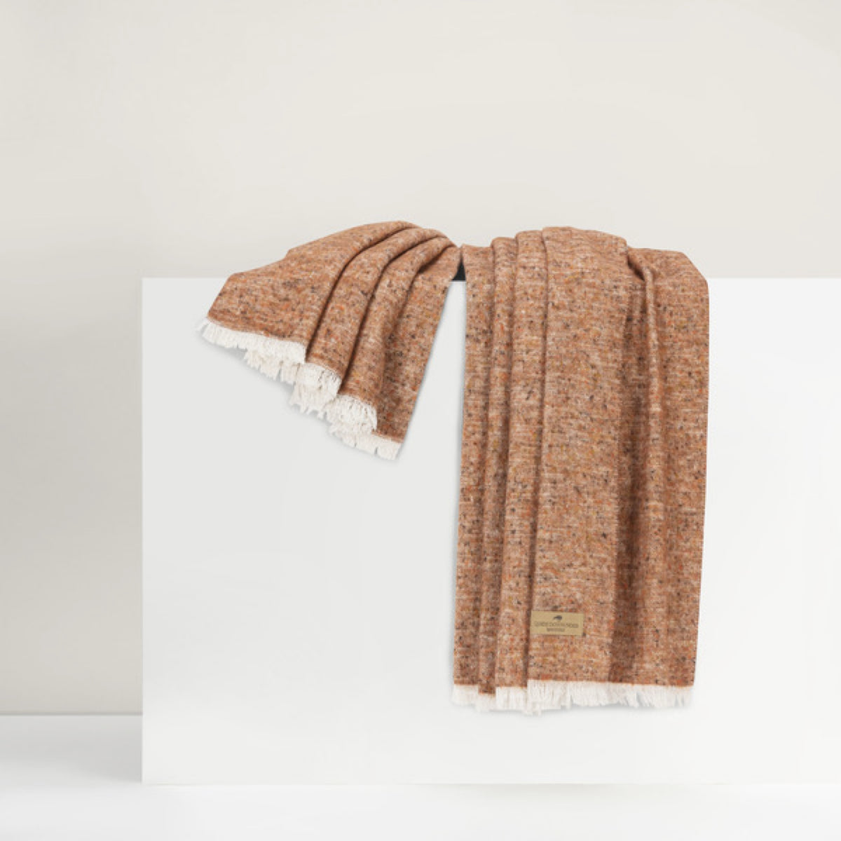 Lands Downunder Cinder Mist Throw in Rust color with fringed edges is draped over a white surface against a neutral background