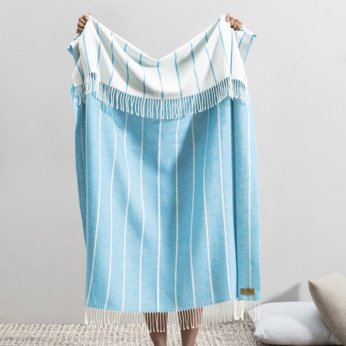 Person holding up an Island Blue Lands Downunder Fiji Stripe Throw, standing on a textured rug in a minimalist room