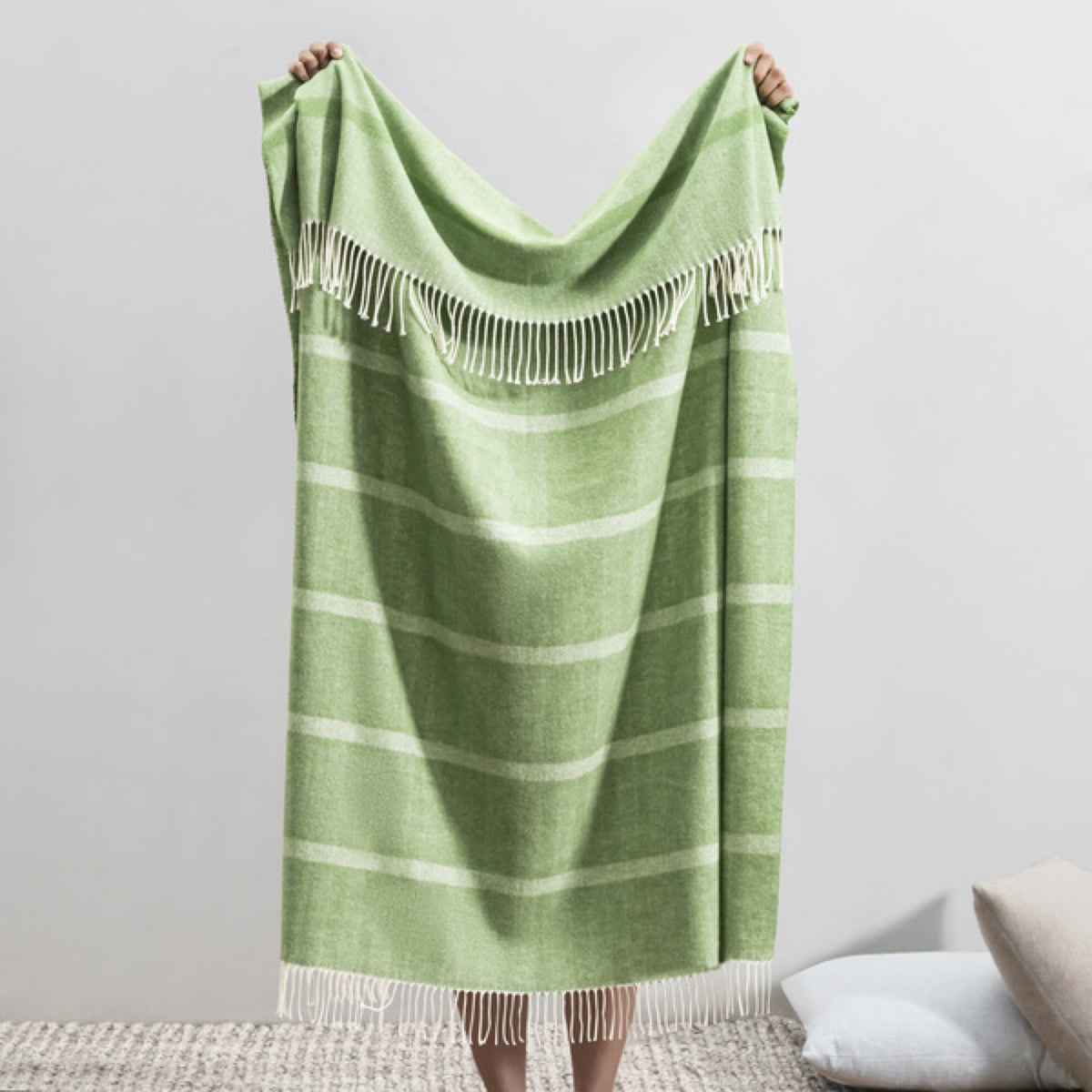 A person displays the Back Lands Downunder Montauk Stripe Herringbone Throws in Meadow color, against a plain backdrop