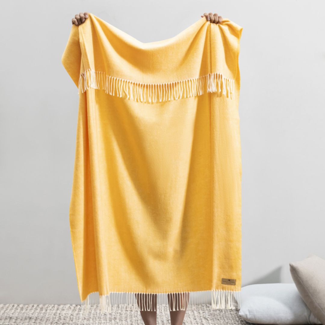 Yellow throw blanket with fringe held by a person against a gray background