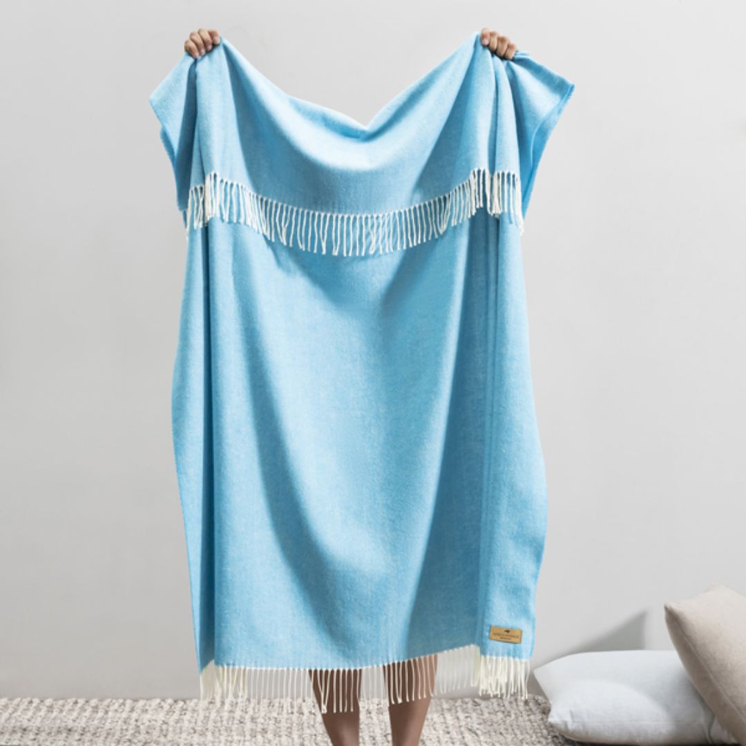 Light blue throw blanket with white fringe held by a person against a neutral background