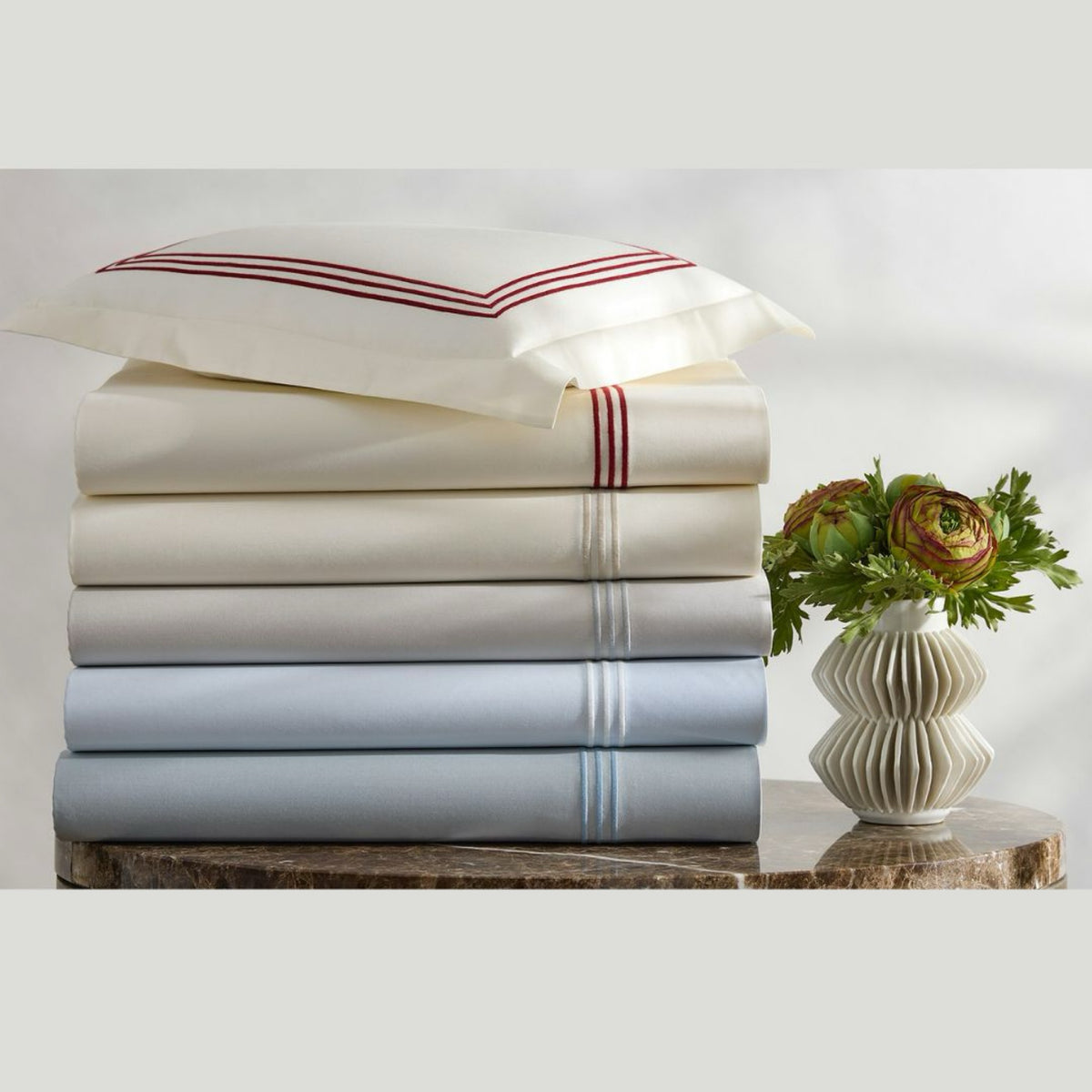 Stack of folded luxury sateen sheets from Matouk Bel Tempo Nocturne Bedding collection, with a sham on top and a vase with flowers on the right