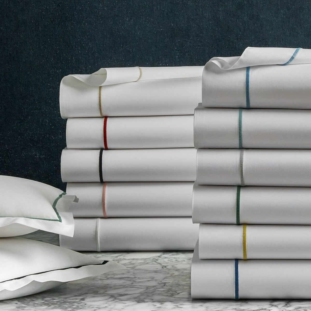 Stacks of folded Bergamo Satin Stitch Bedding in Egyptian cotton percale, displayed on marble against a dark background.