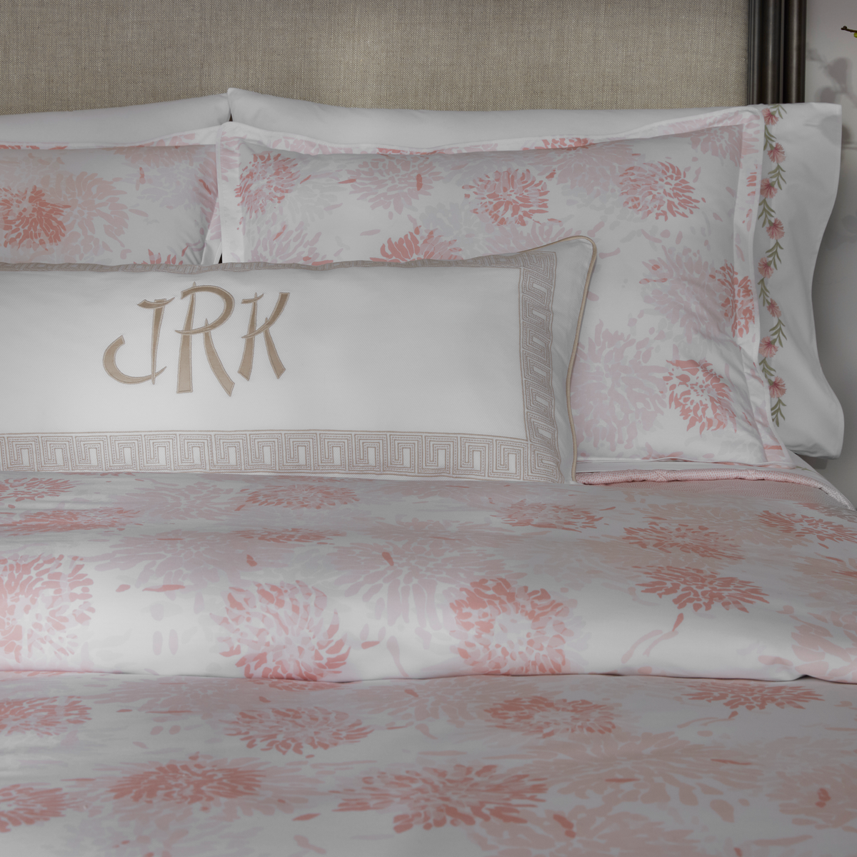 Closeup of a bed with Blush Matouk Chrysanthemum bedding and a rectangular pillow featuring the monogram JRK in beige letters
