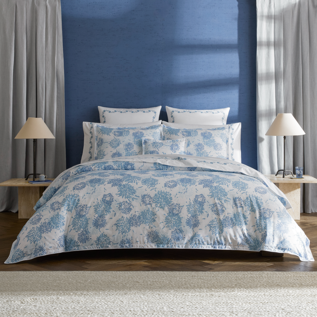 A neatly made bed with Blue Matouk Chrysanthemum luxury bedding, flanked by lamps, stands against a blue wall