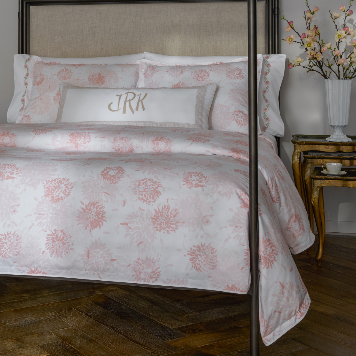 A bed with luxury Blush Matouk Chrysanthemum bedding, a monogrammed pillow, and a vase of flowers in a cozy bedroom