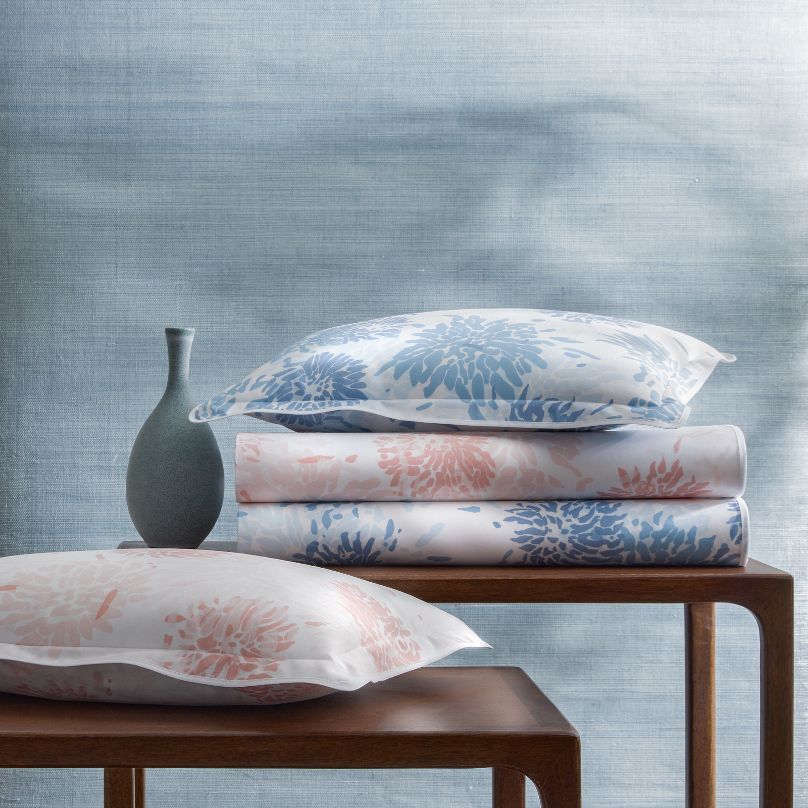 Pillows and folded Matouk Chrysanthemum luxury bedding with floral prints on wooden tables, set against a blue background