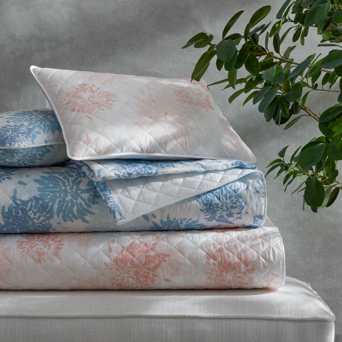 A folded Matouk Chrysanthemum quilt and pillow with blue and blush florals rest on a bench beside a green leafy plant.