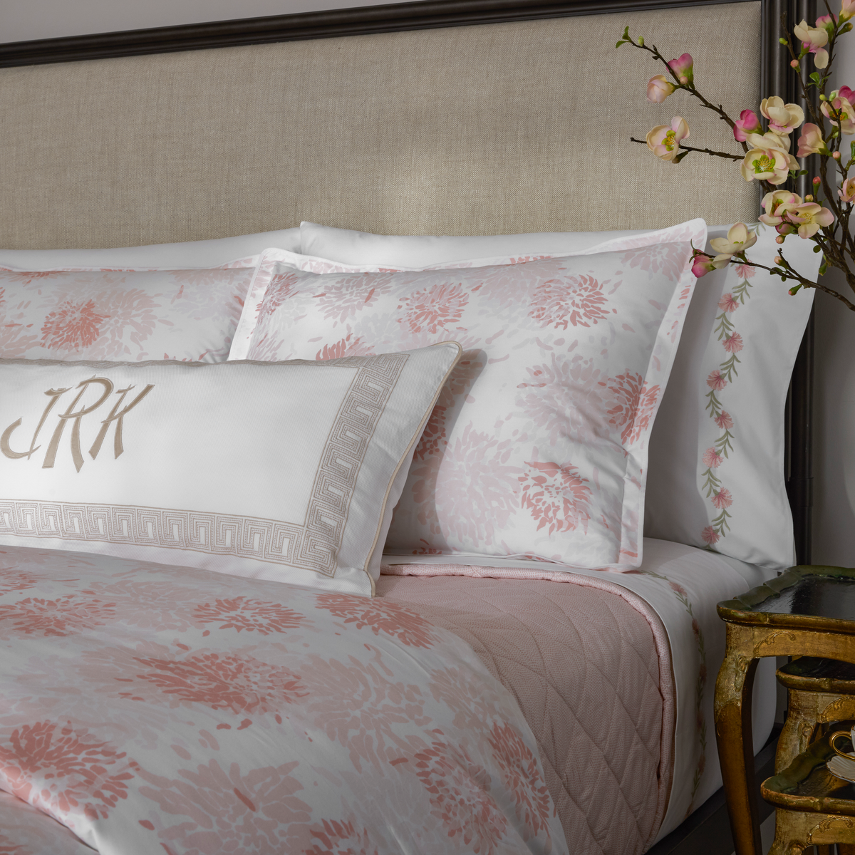A neatly made bed with Blush Matouk Chrysanthemum bedding, embroidered pillows, and flowers on a bedside table.