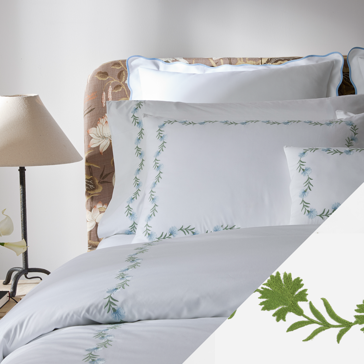 Bed with Matouk Daphne bedding, green floral embroidery, a floral headboard, and beige lampshade on a side table.