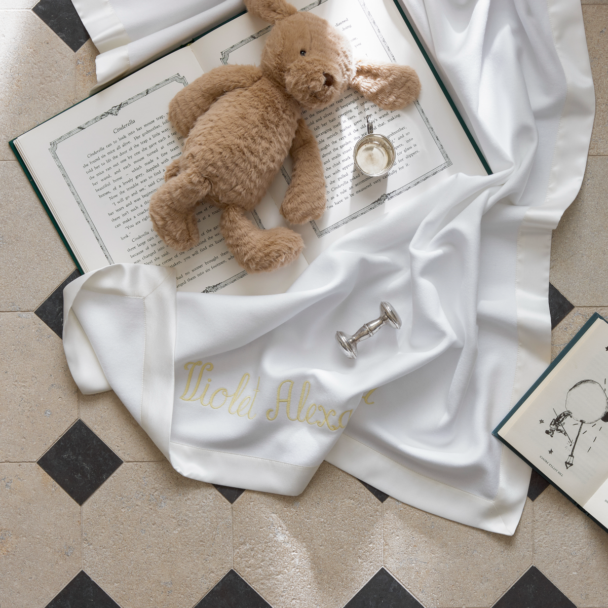 A plush teddy bear, open books, and a soft white Matouk Dream Modal Little Blanket with satin trim rest on a tiled floor