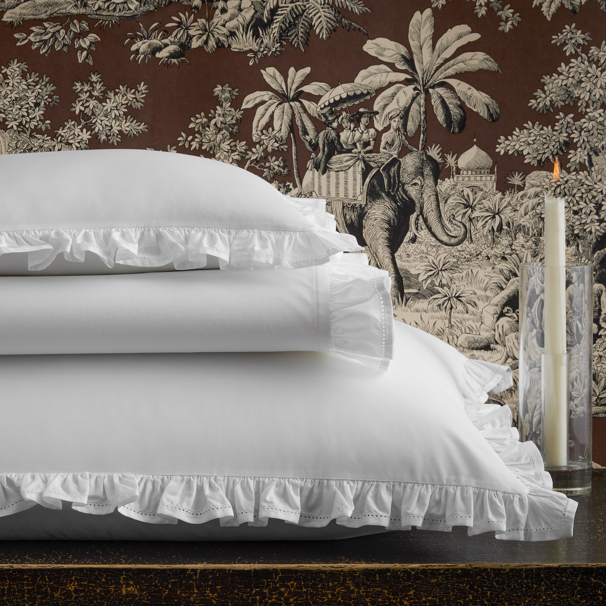 Stacked romantic ruffle bedding from the Matouk Georgia Collection and a lit candle, set against brown jungle-patterned wallpaper.