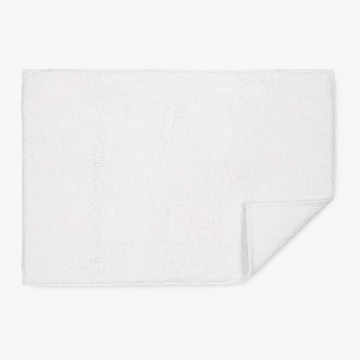 A white rectangular bath mat—crafted like Matouk Giza Towels—has one corner folded, revealing its textured underside.